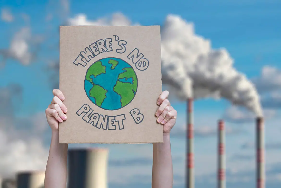 A protest sign is held up high in front of pollution billowing from smokestacks. The protest message is centerd around a blue-green drawing of Earth: There's No Planet B